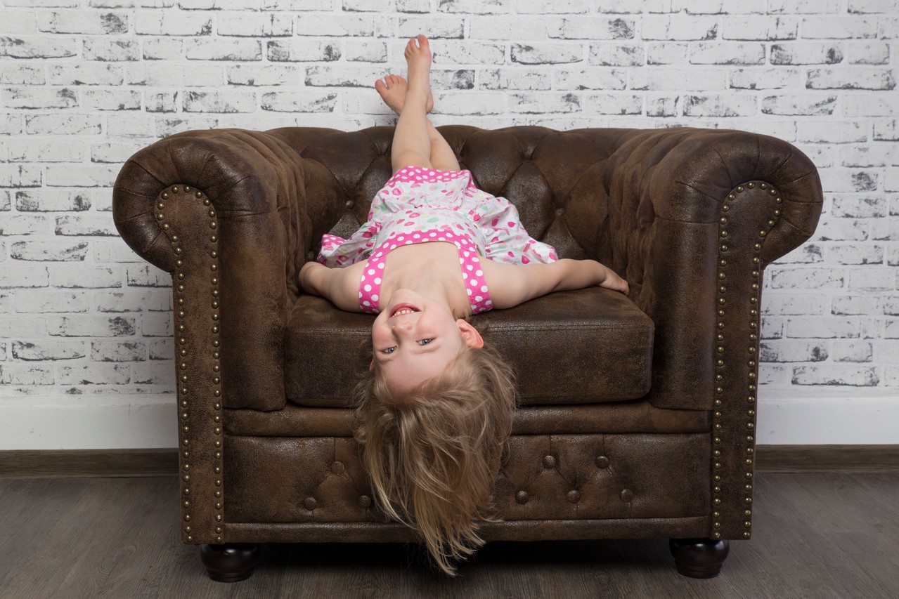 Ein kleines Mädchen sitzt verkehrtherum auf einem Sessel im Fotostudio und lächelt in die Kamera