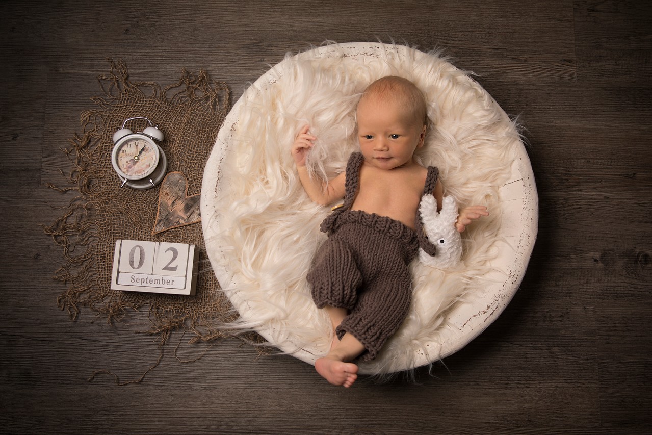 Newbornfotografie - Ein Baby in einer süßesn Strick-Latzhole liegt auf einem Fell in einer Schale